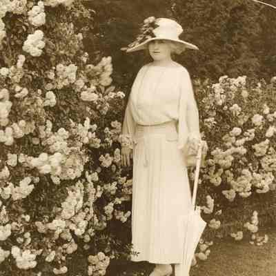 Coe_MC_021: Mai Coe with parasol standing in front of flowering bush
