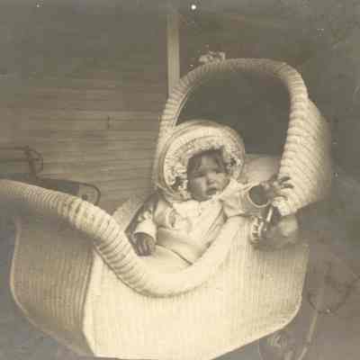 Coe_Natalie_005: Natalie Coe as a baby sitting in wicker high chair and wearing bonnet