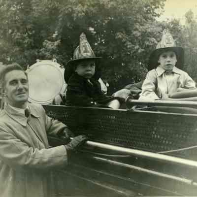 Two young boys with fire hats on sitting in back of fire engine, father standing