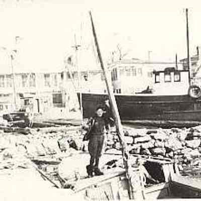 Man on boat tied up at wharf with another boat and houses in background, Stonington CT