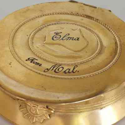 2023.22.1v: Underside of compact which shows two circles the smallest containing a map of Australia, inscribed "Elma", and a boomerang. The larger is inscribed "From Mal."