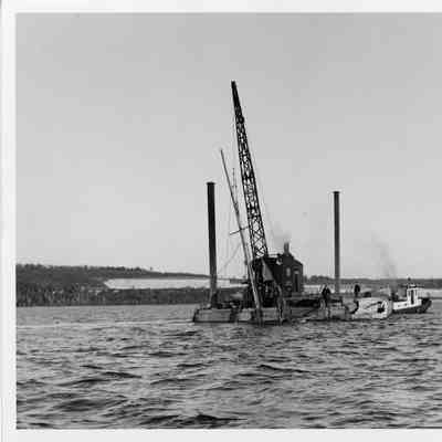 Crane on platform barge holding sailboat being towed by tug boat