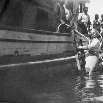 Two people in diving suits partially in water next to boat