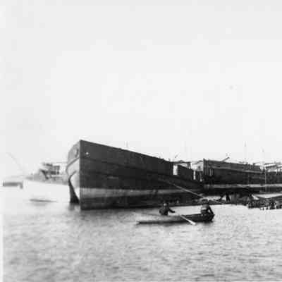 Two men in rowboat next to docked vessels