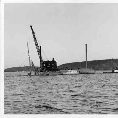 Crane on platform barge holding sailboat being towed by tug boat
