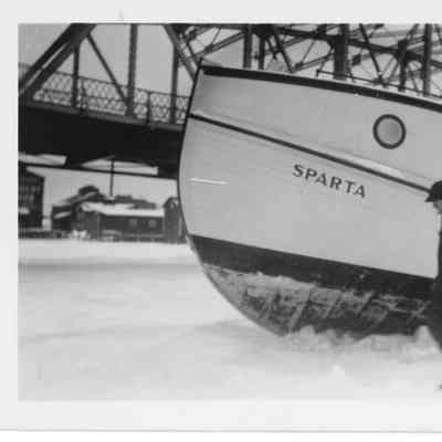 Man walking on ice in front of fishing tug Sparta with Michigan Street bridge in background
