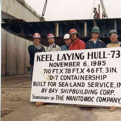 Crew of 7 men standing in back of sign for Keel laying Hull-736 1985