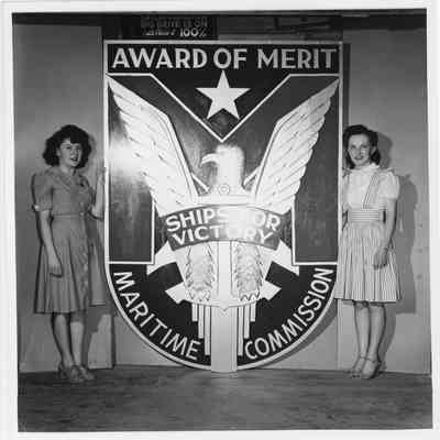 Two unidentified women standing alongside at Award of Merit sign