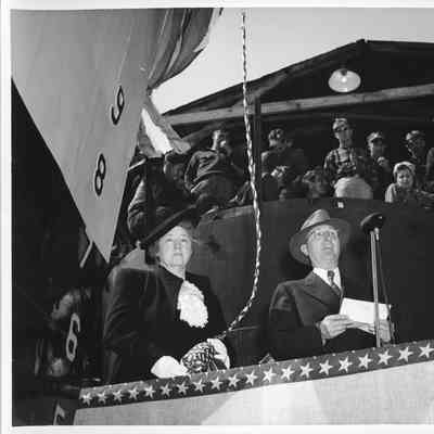 Ship sponsor holding christening bottle next to man giving speech at christening of PC-1177