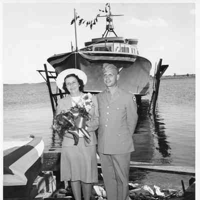Woman holding flowers next to man in uniform with P-571 in background at Peterson Boat Works July 15, 1944