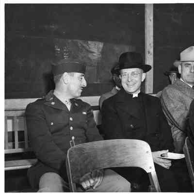 Priest and soldier sitting at ship christening at Peterson Boat Works