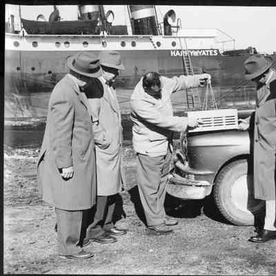 Four men watching another man demonstrate an item on hood of a car