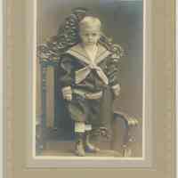 B+W photo of young boy in sailor suit standing on chair, no date (ca. 1900).