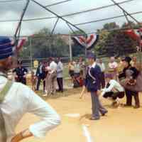 Color photo of a player at bat with catcher and umpire, all in period costume, at Baseball Day Ceremonies, 1976.