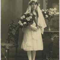 Sepia-tone photo of girl with flowers, catechism, rosary posed in studio with religious interior backdrop, Hoboken, n.d., ca. 1910-1924