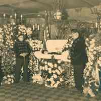 Sepia-tone photo: Funeral bier with an open casket with 2-man honor guard at the Hudsonia Club, Hoboken, no date, ca. 1920.