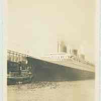 B+W photo of the ocean liner S.S. Normandie at a pier under steam with tugboat at bow. N.d., ca. 1935-1939.