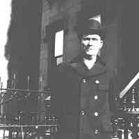 B+W negative image of an unidentified man in topcoat and hat standing near stoop, no place, no date, ca. 1910-20.