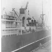 B+W photo of a view amidships of an unknown ship at berth; cargo is being hoisted at center.