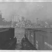 B+W photo of a warship with tugboats in the East River of New York City; car floats in foreground, N.Y., no date, ca. 1940.