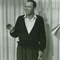 Black-and-white photo of Frank Sinatra in sweater and open shirt collar with cigarette, no place, no date, ca. 1955.