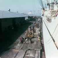 Color photo of loose cargo on pier next to freighter, attributed to Hoboken, n.d., ca. 1970s.