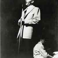 Black-and-white photo of a young Frank Sinatra with the Tommy Dorsey Band singing onstage at microphone, no place, no date, ca. 1940-42.