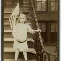 Sepia-tone photo of a young girl holding flag on stick standing on front steps of apartment building, Hoboken? N.d., ca. 1910-1920.