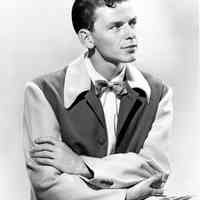 Black-and-white photo of a young Frank Sinatra seated in a lounging jacket & bowtie, no place, no date, ca. 1939-1943.