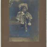 Sepia-tone photo of an unidentified young girl, ca. 3 years old, posed next to a chair in a studio, Hoboken?, no date, ca. 1900-1910.