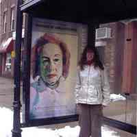 Color photograph of Laura Alexander standing in bus shelter with panel advertising exhibition of her paintings, Hoboken, ca. February-March 2005.