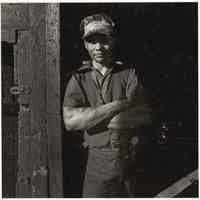 B+W photo of a man dressed in workclothes leaning against an aged wood wall, Hoboken, no date, [1976].