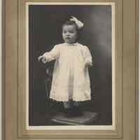Sepia-tone photo of an unidentified baby, ca. 1-2 years old, posed standing on a chair in a studio, New York City, no date, ca. 1900-1910.