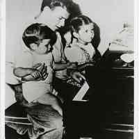 B+W photo of Frank Sinatra holding Frank, Jr. seated at a piano with daughter Nancy, n.p., n.d., ca. 1945.