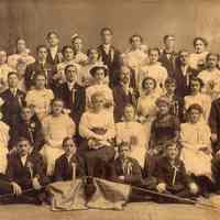 Sepia-tone group photo of three adults with a large group of children in formal dress posed in photographer