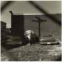 B+W photo of two cars and a boat in a fenced-in lot, Hoboken, no date, [1976].