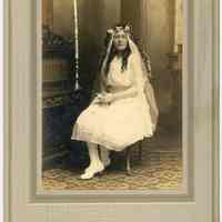Sepia-tone photo portrait of young woman in confirmation dress with flower headdress posed in photographer