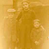 Digital image of photo of a girl with two boys posed outdoors near rough planked wall, no place (Hoboken?), no date, circa 1870