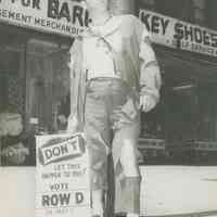 B+W photo of a man posed standing at astreet curb dressed in ragged clothing for a political campaign, Hoboken, no date, ca. 1953.