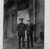 B+W photo of a policeman and man with cap posing in front of brick building, Hoboken shipyard, no date, ca. 1940.