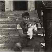 B+W photo of a man seated on a front stoop holding a very young baby, Hoboken, no date, [1976].