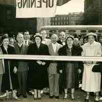 B+W photo of Nathan Marcus, his wife and store staff posing in front of the ribbon at the Grand Opening of Marcus Jewelers, Hoboken, April 3, 1954.