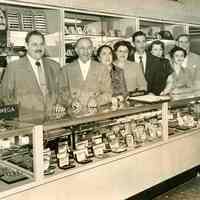 B+W photo of Nathan Marcus, his wife and store staff posing behind a counter at the Grand Opening of Marcus Jewelers, Hoboken, April 3, 1954.