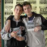 Color photo of two boys posed with Burczy exhibit postcard in front of museum display case, Hoboken, May 21, 2006.