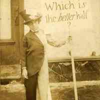 Image: sepia-tone photograph of a girl in costume holding a sign "Which is the better half?" no date, ca. 1910-20.