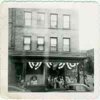 B+W photo of a decorated Hoboken storefront with children wearing ceremonial sashes around the doorway, Hoboken, no date, ca. 1955.