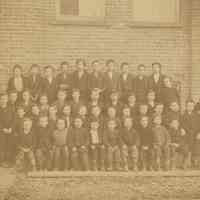 Sepia-tone group photo of schoolboys with a nun and priest outside what is probably the school building, no place, no date, ca. 1900.