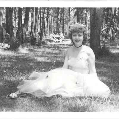 Pageant queen Audrey Moe sitting on grass while wearing a white dress, a small picnic table visible in the trees behind her