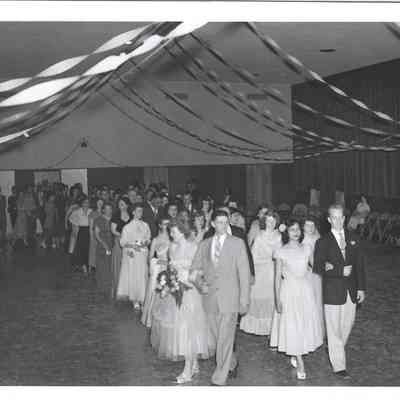 Two lines of young women arm-in-arm with adult men walking under streamers