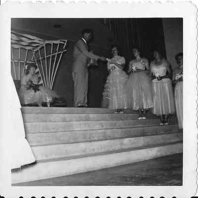Pageant princesses on stairs receive something from pagent emcee under watchful eye of pageant queen